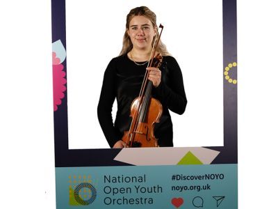 A girl with long blonde hair and wearing a shiny black dress holds a violin. She stands in between a customised National Open Youth Orchestra Instagram frame. She is smiling.